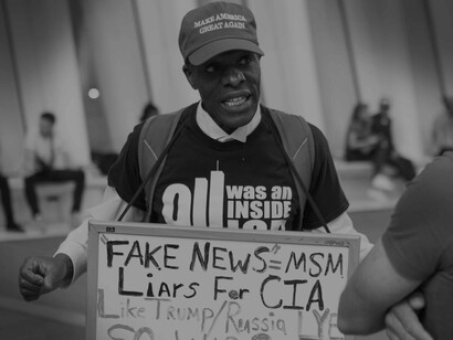 A man carrying a bulletin board, and wearing a MAGMA cap is spreading his truth about how the news is spreading a false reality