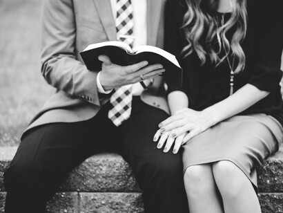 A greyscale image of a couple in formal attire, reading a book together in a garden