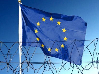 The European Union flag waves in the cloudy sky outside the prison, just beyond the barbed wire — a stark symbol of border security