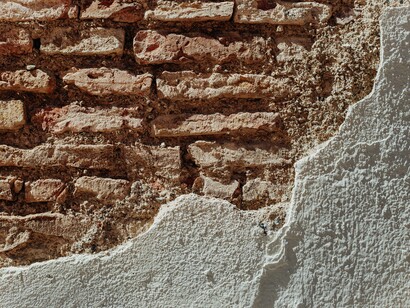 Un primo piano di un muro danneggiato in cui una parte dell'intonaco bianco si è staccata, lasciando scoperti i mattoni rossi sottostanti