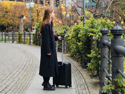 A young woman in a coat walks thoughtfully through a city street with a black suitcase, reflecting the experience of moving abroad and starting a new life