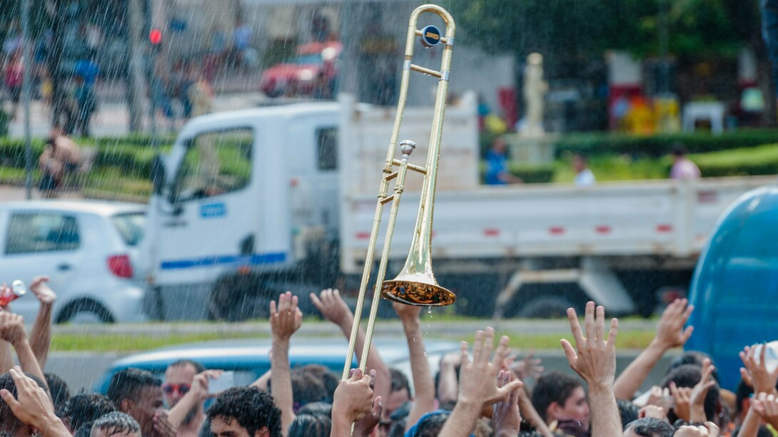 Aglomeração de pessoas com um trombone em destaque entre a multidão. A imagem remete à expressão “botar a boca no trombone”, usada quando alguém torna público aquilo que pensa, denuncia uma situação ou decide falar abertamente sobre algo — um exemplo de como o sentido figurado transforma uma expressão cotidiana