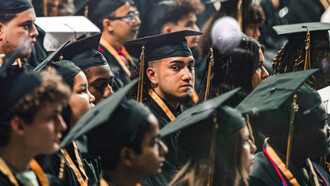 Un joven apelando a la cámara con mirada seria en el día de su graduación, junto al resto de sus compañeros