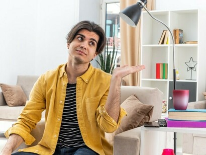 A young man in casual clothes sits on a chair, looking to the side with a pensive expression, his hand resting on his chin