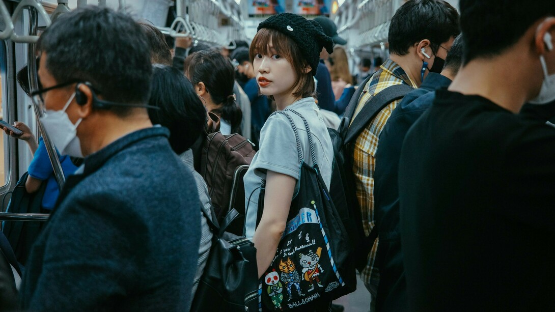 A young Korean woman stands on a subway train, gazing directly at the camera, surrounded by other commuters
