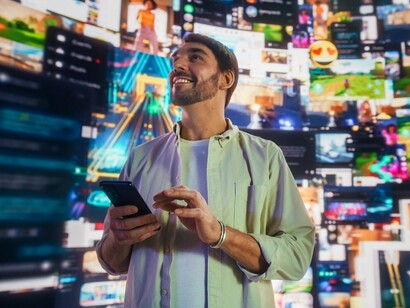 Um homem sorri enquanto segura o smartphone diante de um mosaico de imagens e conteúdos digitais. Entre tantas possibilidades, o algoritmo procura identificar aquilo que pode prender nossa atenção