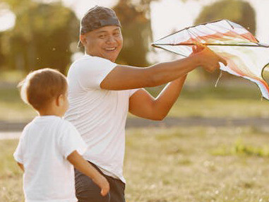 Father smiles at his son while flying a kite, appreciating the beauty of the present moment