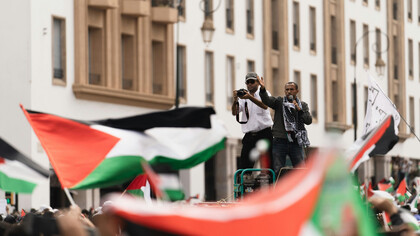 A man holding a microphone speaks to a crowd, as several Palestinian flags wave