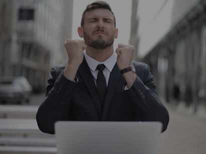 A man in a sleek black suit sits on a chair amidst towering urban buildings