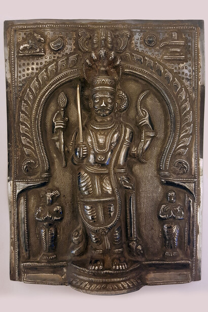 Vīrabhadra con gli occhi risaltanti e i tre segni orizzontali simbolo del dio Śiva sulla fronte. India meridionale. Sbalzo su lega di argento. Cm. 20,6 x 15,6