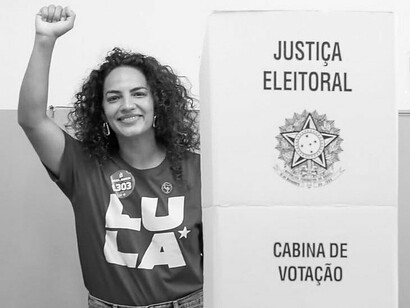 Uma eleitora ergue o punho diante da cabine de votação da Justiça Eleitoral, vestindo uma camiseta de apoio a Lula. O gesto traduz a dimensão política do voto como expressão de convicções, participação e escolha