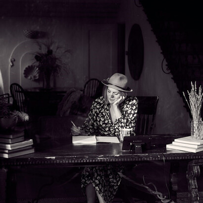 Captured in classic black and white, a talented woman author donning a stylish hat sits on a chair, engrossed in her writing, her pen gracefully gliding across the page, an embodiment of creative focus and elegance