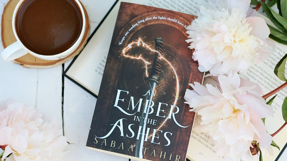 A cup of coffee and a copy of An Ember in the Ashes on a cozy wooden table