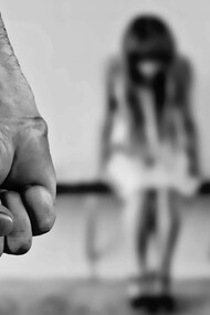 A man stands with his fists clenched in anger, while a woman sits on the ground in front of him, fearful and covering her face