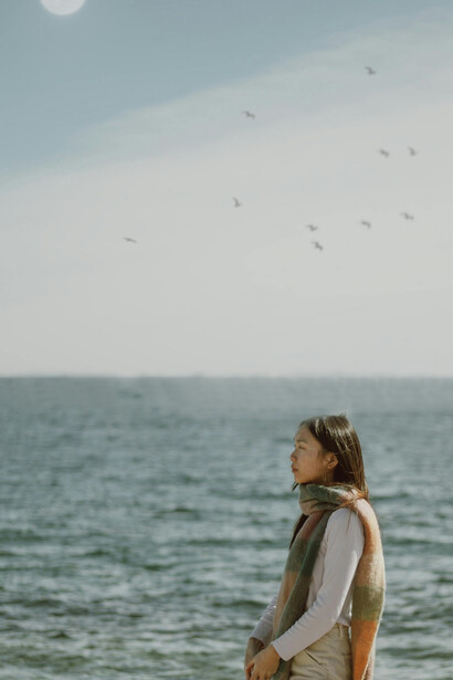 An Asian woman with long hair standing by the sea, calmly observing the waves, evoking a quiet sense of solitude and loneliness