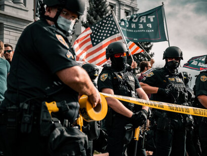 A man in a black police uniform stands amidst a crowd during a daytime protest in the USA, symbolizing the growing tension and division in society