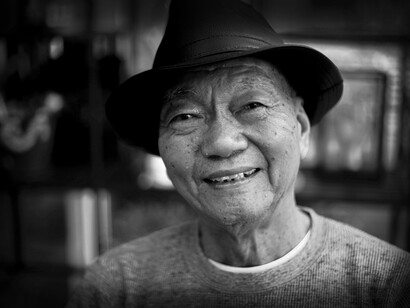 An elderly Asian man smiles wearing a hat