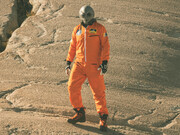 An alien humanoid figure in an orange jumpsuit looks at the camera