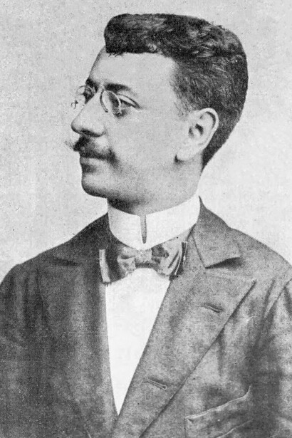 Olavo Bilac em um retrato de perfil, usando terno, gravata e óculos, em uma fotografia em preto e branco. A imagem remete ao poeta parnasiano cuja obra exemplifica a atenção ao rigor formal e ao uso cuidadoso das palavras, mostrando como a escolha e a organização da linguagem também podem funcionar como recursos de construção de sentido e persuasão