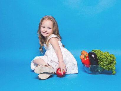 A young girl holding vegetables in front of a blue wall, learning about ingredients and making healthy food choices