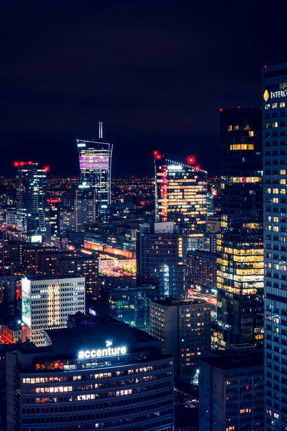 A cityscape of Warsaw at night, with a Ferris wheel glowing in the background, showcasing the urban roads and vibrant city atmosphere