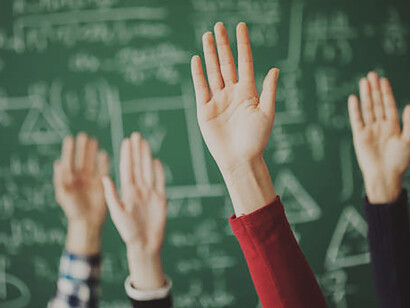 Students raising their hands against a green board in a classroom provides a counterpoint to the principles endorsed by Rousseau