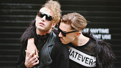 Dois homens abraçados com visual inspirado na moda punk e gótica. A Versace Spring–Summer 2023 trouxe elementos dramáticos e góticos, fazendo referências sacras com véus, crucifixos e muitas texturas