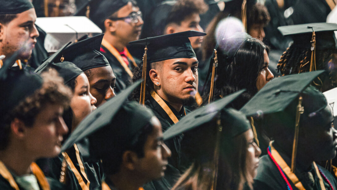 Un joven apelando a la cámara con mirada seria en el día de su graduación, junto al resto de sus compañeros