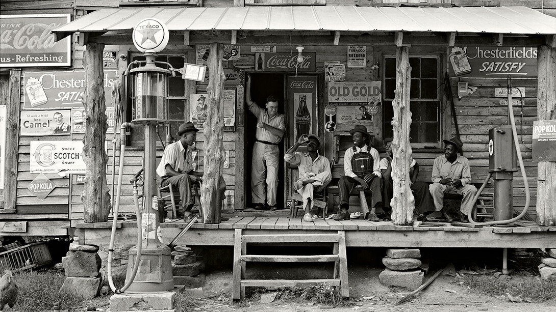 Dorothea Lange, Negozio di campagna su strada sterrata. Domenica pomeriggio. Si noti la pompa del cherosene a destra e quella della benzina a sinistra. I pali di legno grezzi e non rifiniti sono stati usati come sostegno per il tetto della veranda. Uomini neri sono seduti sulla veranda. Il fratello del proprietario del negozio è in piedi sull'ingresso - Gordonton, North Carolina, 1939. Per gentile concessione di Anna Defrancesco