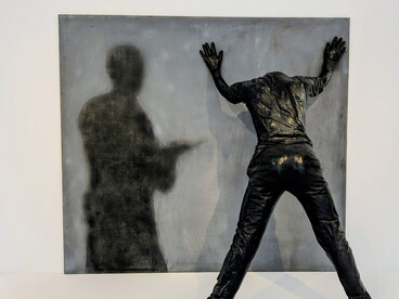 Rafael Canogar (b. 1935). The artwork in the image is titled El Arresto II (The Arrest II) (1972). Canogar is a significant Spanish artist known for co-founding the El Paso group and for his work in both informal abstraction and figurative art with socio-political themes, such as police brutality. This piece is from Canogar's second figurative period. It breaks the barrier between painting and sculpture to create a 3D effect, focusing on themes of dehumanization, repression, and social critique. The sculpture of the figure against the wall with hands up is depicted alongside a painted shadow figure holding what appears to be a gun. Image ©MACA/the Author 2026