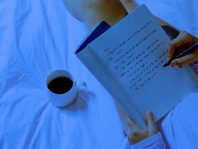 A woman, laying on her bed, busy handwriting on her journal, while having a cup of coffee. The most efficient way forward for writers is to accept the non-human competition, assess it, and use it to their advantage
