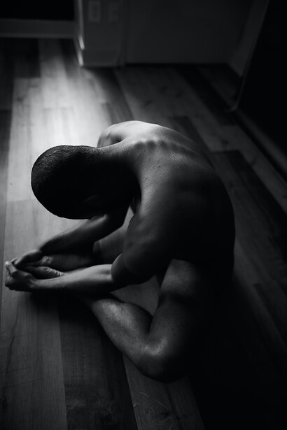 In a mesmerizing grayscale image, a man kneels on the floor, radiating a captivating blend of beauty, strength, and vulnerability