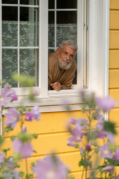 O homem apoia-se no peitoril enquanto observa o movimento além da casa. Flores desfocadas ocupam o primeiro plano, enquanto a figura permanece recolhida à janela. Às vezes, o vazio instala-se devagar, como uma visita habitual. A vida continua a passar mesmo quando alguém decide parar para a observar