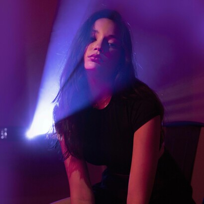 Donna con lunghi capelli scuri, illuminata da un fascio di luce alle spalle che crea forti contrasti viola scuro sullo sfondo