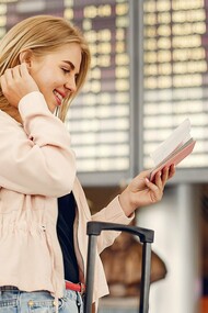 A smiling traveler checks her passport in front of an airport departures board, reflecting the article’s focus on mindful preparation and a stress-free travel mindset