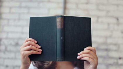 Mulher com livro aberto em frente ao rosto. Primeiro de tudo, vamos deixar claro: sim, qualquer leitura ajuda! E não, isso não é uma desculpa para se entupir de revistas de fofoca e achar que está tudo bem (apesar de que, se isso te faz feliz, quem sou eu para julgar?). O ponto é que ler, independentemente do que seja, ativa o cérebro, melhora o vocabulário e a compreensão, além de nos transportar para outros mundos e nos fazer enxergar as coisas de outra maneira