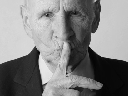 A senior citizen has his finger to his lips taunting people to stay silent