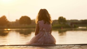 A young girl sitting alone beside the water at sunset, representing the loneliness that can begin with the fear of being left