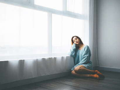 A woman sits on the floor beside a window, her posture reflecting a sense of melancholy