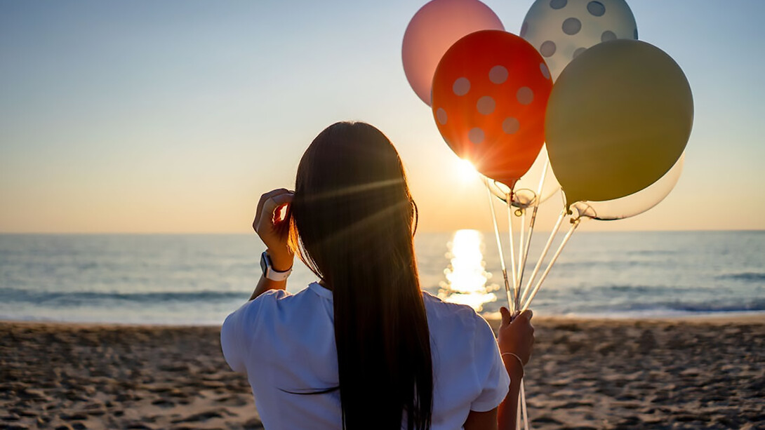 A woman standing on the beach, holding colorful balloons, with her back to the camera