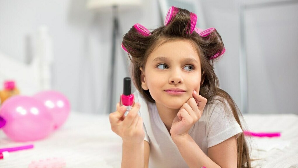 Preschool-aged girl lying on a white blanket with hair curler, holding nail polish 