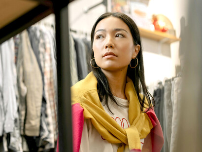 A woman in a white long-sleeve shirt and blue denim jeans browses through clothing racks at a secondhand store