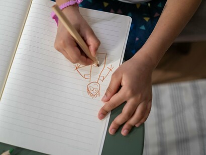 A menina continua desenhando sobre a folha, com o lápis conduzindo a construção de uma imagem ainda em formação. A cena remete à infância como espaço de aprendizagem, quando representar uma família também significa descobrir quais presenças fazem parte dela — e quais permanecem ausentes