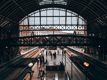 Travellers waiting beneath towering steel arches, symbolising the uncertain beginnings that accompany every journey toward a new home