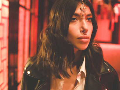 An Indian woman wearing a leather jacket, featuring a septum piercing and traditional maang tikka, looks thoughtfully afar