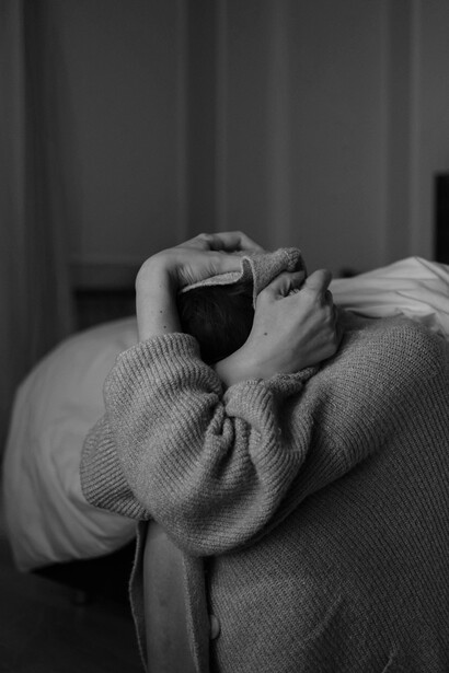 A woman sits wrapped in a blanket, her hand on her head, conveying anxiety and distress