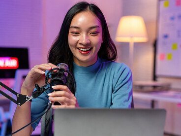 A young Asian content creator hosts a podcast from her home studio, engaging with her audience through a microphone and laptop setup
