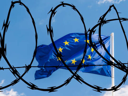 eyond the barbed wire of the prison, the European Union flag flutters under a cloudy sky, highlighting the tension of border control