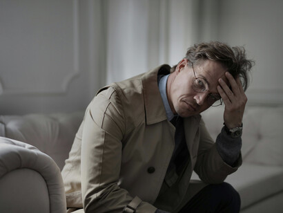 A mature man sits alone on a couch, thinking in a dimly lit living roo