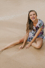 A cheerful and radiant woman sits on the sandy beach, wearing a smile that brightens the surroundings